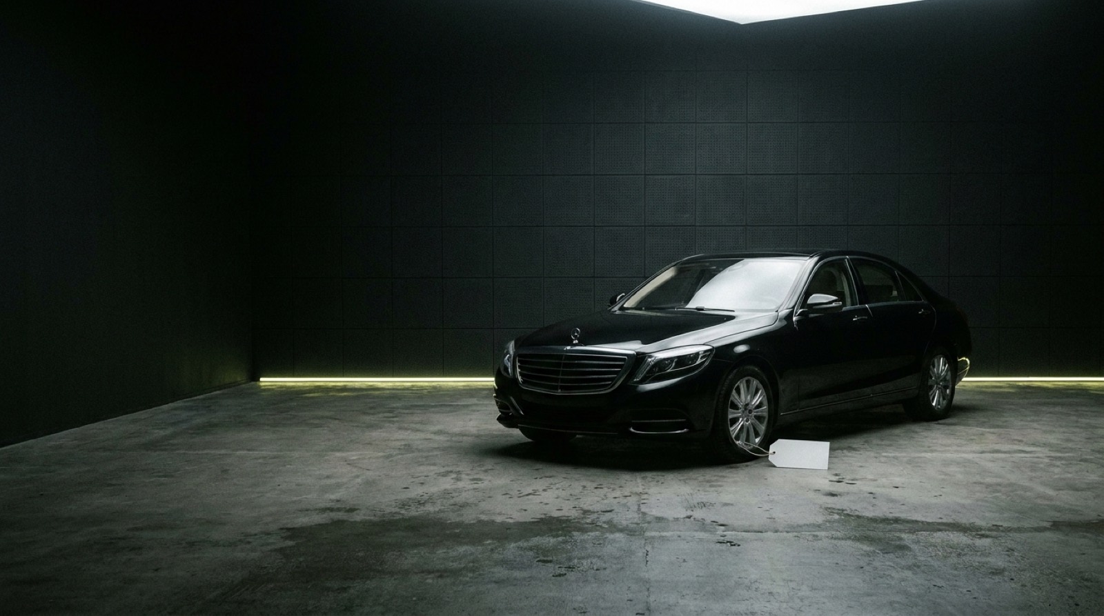 A black luxury executive sedan alone in a dark concrete room, a blank price tag on the floor beside it.